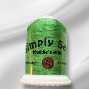 Peut inclure: Une bobine de fil à broder vert avec le texte "Simply Sew Maddie's Attic" et "Polyester Embroidery" sur l'étiquette. Un bouton marron à quatre trous est sur l'étiquette.