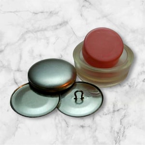May include: A set of three silver metal buttons with a loop on one button. A pink plastic button is shown in a clear plastic container.