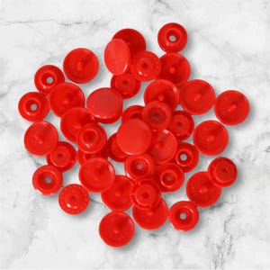 Puede incluir: Un montón de botones a presión de plástico rojo. Los botones son de varios tamaños y formas, y algunos tienen un centro elevado.