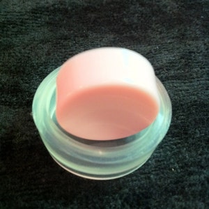 Pode incluir: Uma barra de sabão rosa em um anel de plástico transparente. O sabão é redondo e tem uma superfície lisa.