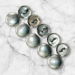 May include: A set of nine silver-toned buttons with a shiny finish. The buttons are round and have a raised center. They are arranged in a row on a white marble surface.