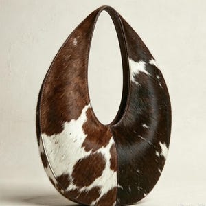 May include: A crescent-shaped hobo bag featuring a brown and white cowhide pattern with a textured fur finish. The bag has a single integrated handle and dark brown leather trim along the edges, creating a distinct oval silhouette.