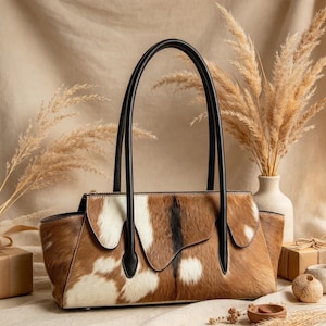 May include: A handbag featuring a brown and white cowhide pattern with a textured fur finish. The bag has two tall, smooth black leather handles and a top zipper closure. The design includes a flap detail on the front and a structured base.