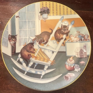 May include: A round decorative ceramic plate with a gold rim featuring a detailed illustration of four brown tabby kittens playing on and around a white wooden rocking chair in a room filled with various toys, including a doll and a toy house.
