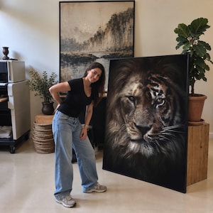 May include: A large rectangular framed wall art piece featuring a split-face portrait of a lion and a tiger. The artwork displays the detailed fur textures and intense gaze of both animals merged into one cohesive image with a dark background.