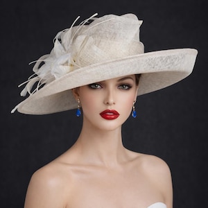 May include: A cream-coloured wide-brimmed hat with a layered crown and feather detailing. The image also features blue teardrop earrings and red lipstick against a dark grey backdrop.