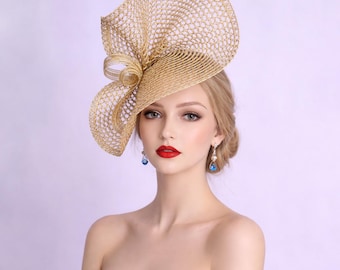 May include: A gold-colored, open-weave hat with a large bow detail. The hat is worn by a person with blonde hair and red lipstick. The hat appears to be made of a woven material, and the overall style is elegant and formal.