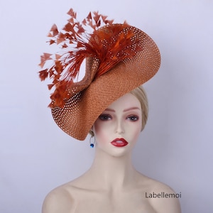 May include: A large, brown, straw hat with a wide brim and a large, orange feather accent. The hat is tilted at an angle and is worn by a mannequin.