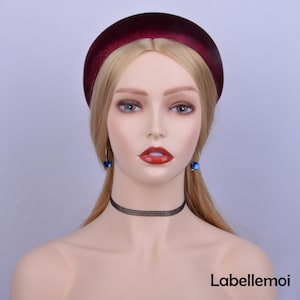May include: A burgundy velvet headband with a padded design. The headband is worn by a mannequin with blonde hair and blue earrings. The mannequin is wearing a black choker necklace.