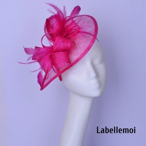 Op de afbeelding: Een felroze fascinator met een gebogen, opengewerkte rand en een decoratieve strik. De hoofdtooi is versierd met veren en een haarband. De tekst "Labellemoi" is rechtsonder zichtbaar.