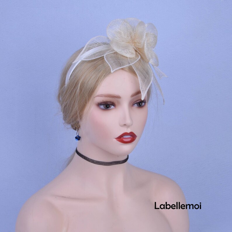 May include: A white headband with a large, ivory-colored flower made of netting. The headband is worn on a mannequin with blonde hair.