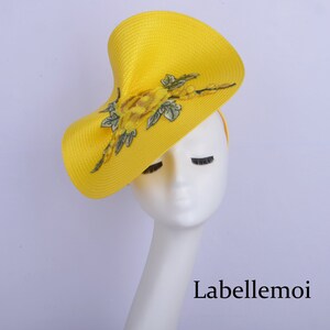 May include: A bright yellow hat with a large, asymmetrical brim and a floral design. The hat is made of straw and has a delicate, embroidered floral pattern in shades of yellow and green.