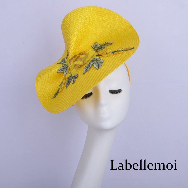 May include: A bright yellow hat with a large, asymmetrical brim and a floral design. The hat is made of straw and has a delicate, embroidered floral pattern in shades of yellow and green.