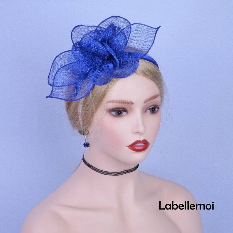 May include: A blue flower headband with a large, layered flower made of netting. The headband is worn by a mannequin with blonde hair.