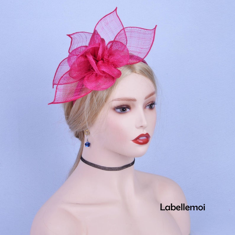 May include: A pink flower hair clip with a large, ruffled flower and a delicate mesh veil. The clip is worn on a mannequin with blonde hair.