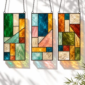 May include: Three rectangular stained glass panels hanging from black metal chains. Each panel features a geometric arrangement of multicolored textured glass pieces in shades of green, blue, yellow, orange, and red, framed in black metal.