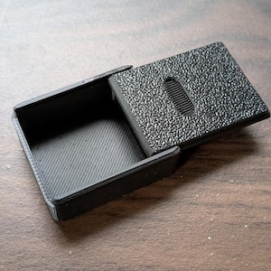 May include: A small, rectangular black plastic container with a sliding lid. The base features a diagonal textured pattern, while the top surface of the sliding cover has a rough, pebbled texture and a central oval-shaped grooved grip.