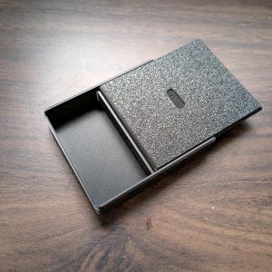 May include: A black rectangular sliding case with a textured top surface and a smooth, open compartment. The lid features a small, centered oval indentation for grip, and the interior base has a fine, diagonal lined pattern.