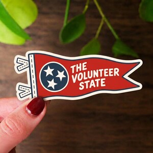 May include: A red, white, and blue pennant-shaped sticker featuring a dark blue circle with three white stars. The text THE VOLUNTEER STATE is printed in white capital letters on the red background. The sticker has a white border and two ties.