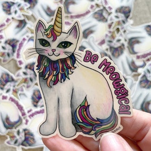 May include: A white cat with a rainbow mane and a unicorn horn, sitting with its tail curled around its legs. The cat has green eyes and a pink nose. The background is a white surface with the text "Be Meowsical" in pink letters.