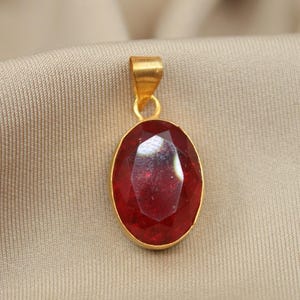 May include: An oval-shaped pendant featuring a faceted deep red gemstone set in a polished gold-toned metal bezel. The pendant includes a matching gold-toned metal bail at the top, designed for attachment to a necklace chain or cord.