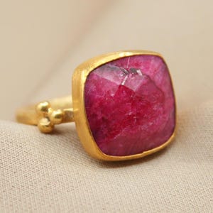May include: A gold ring featuring a large, square-cut, faceted deep pink gemstone set in a bezel. The band has a matte gold finish and includes a small decorative cluster of three tiny gold spheres where the band meets the setting.