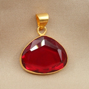 May include: A teardrop-shaped pendant featuring a deep red, faceted gemstone set in a gold-toned metal frame. The pendant includes a smooth, gold-toned bail at the top for attachment to a chain, showcasing a polished finish and reflective surfaces.