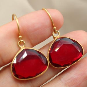 May include: A pair of drop earrings featuring faceted, deep red, pear-shaped stones set in a matte gold-toned metal frame. Each earring hangs from a simple gold-toned hook wire, showcasing a polished finish on the stones and a textured gold setting.