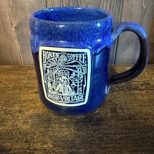 May include: A deep blue ceramic coffee mug with a glossy, mottled finish and a large handle. A rectangular white label on the front features a black graphic of a skeleton in a lab coat surrounded by scientific equipment, with the text BONES COFFEE CO. HIGH VOLTAGE.