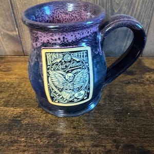 May include: A ceramic mug with a dark blue and purple speckled glaze featuring a rectangular label that reads Bones Coffee Red Velvet with an illustration of a winged skeleton holding a heart that says Be Mine in black and white ink.