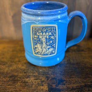 May include: A bright blue ceramic mug with a handle, featuring a rectangular cream-colored label on the front. The label displays the text "Bones Coffee Co." and "Shark Bite" above an illustration of a shark biting a skeleton in the ocean.
