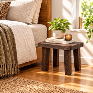 May include: A dark brown wooden side table with four sturdy legs. On top sits a potted green plant in a speckled ceramic pot, a stack of two white books with the text KINFOLK on the spine, and a small amber glass candle holder with a lit candle.