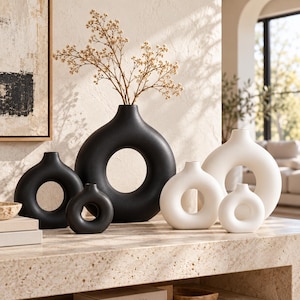 May include: A collection of five circular, donut-shaped vases in matte black and matte white finishes. The vases feature a central hollow opening and a narrow neck. One large black vase contains a sprig of dried baby's breath flowers.