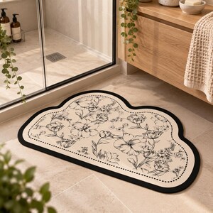 May include: A cloud-shaped bathroom floor mat featuring a cream-colored surface with a black floral line art pattern. The mat has a thick black border and a dotted inner trim, creating a distinct outline against the tiled floor.