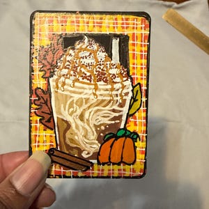 Könnte beinhalten: Eine rechteckige Holzplakette mit einer handgemalten Illustration eines Kürbisgewürz-Lattes mit Schlagsahne, Karamellsoße und Zimtstangen. Der Hintergrund zeigt ein gelb-orangefarbenes Karomuster mit herbstlichen Blättern.