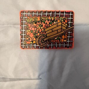 Può includere: Una targa rettangolare in legno con un'illustrazione dipinta a mano di una carriola marrone piena di zucche arancioni. La scena è incorniciata da un motivo a griglia bianco e nero con un bordo arancione brillante attorno.