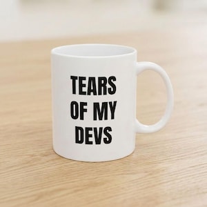 Könnte beinhalten: Ein weißer Keramikbecher mit C-förmigem Henkel, der auf der Vorderseite den schwarzen Schriftzug "TEARS OF MY DEVS" in einer fetten, serifenlosen Schrift trägt. Der Becher hat einen glatten, zylindrischen Körper und einen Standardrand.