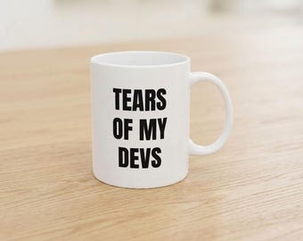 May include: A white ceramic mug with a C-shaped handle featuring the black text "TEARS OF MY DEVS" printed in a bold, sans-serif font on the front. The mug has a smooth, cylindrical body and a standard rim.