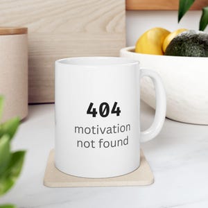 Könnte beinhalten: Ein weißer Keramikbecher mit dem schwarzen Text 404 und dem grauen Text motivation not found, der mittig auf der Vorderseite steht. Der Becher hat eine zylindrische Form mit einem geschwungenen Henkel an der rechten Seite.
