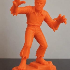 May include: A bright orange plastic figurine of a werewolf standing on a rocky base. The figure is depicted in a crouched, aggressive pose with arms raised, hands clawed, and wearing a collared shirt and pants with tattered, jagged hems.
