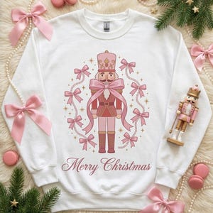Op de afbeelding: Een wit sweatshirt met ronde hals en een centrale afbeelding van een roze notenkraker, omringd door een cirkel van roze strikken en gouden sterren. De tekst Merry Christmas staat in een cursief roze lettertype onder de notenkraker.