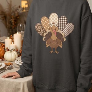 Op de afbeelding: Een donkergrijs sweatshirt met ronde hals en een centrale afbeelding van een kalkoen met een staart in verschillende patronen. De kalkoen heeft een klein wit strikje op het hoofd en de staartveren tonen diverse prints zoals luipaard, stippen, ruitjes en strepen.