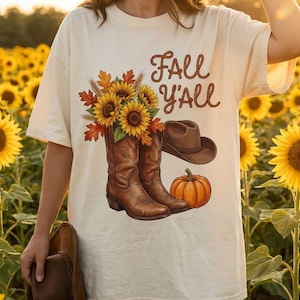 Op de afbeelding: Een crèmekleurig t-shirt met een grafische print van bruine cowboylaarzen gevuld met zonnebloemen en herfstbladeren, een bruine cowboyhoed, een oranje pompoen en de bruine tekst "FALL Y'ALL" in een touwachtig lettertype.