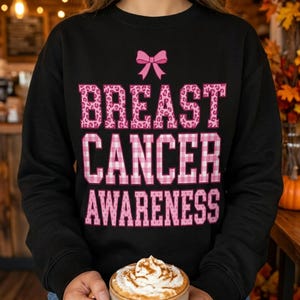 Op de afbeelding: Een zwart sweatshirt met lange mouwen en een roze strikgrafiek boven de tekst BREAST CANCER AWARENESS. Het woord BREAST heeft een roze luipaardprint, CANCER een roze en wit gingham-patroon en AWARENESS een roze stippenpatroon.