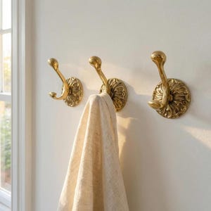 Peut inclure: Trois patères murales en laiton poli avec des plaques arrière circulaires ornées de détails floraux complexes. Chaque crochet présente un design incurvé à double pointe. Un tissu texturé de couleur claire est suspendu au crochet central.