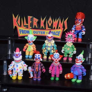 Może przedstawiać: Kolekcja ośmiu figurek akcji Killer Klowns from Outer Space ustawionych na stopniowanym stojaku. Figurki mają kolorowe, szczegółowe kostiumy klaunów, pomalowane twarze i różne akcesoria, a za nimi znajduje się logo filmu.