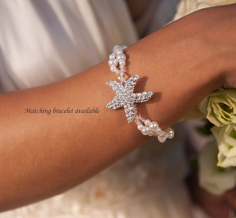 May include: A silver bracelet with a starfish charm. The starfish is encrusted with small, sparkling crystals. The bracelet also features strands of small, white pearls. The text "Matching bracelet available" is visible.