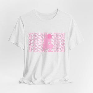 May include: A white short-sleeved t-shirt featuring a pink graphic print on the chest. The design consists of the word STANDARDS repeated in six horizontal lines with a silhouette of a person crouching in the center of the text.