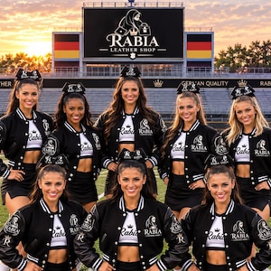 Puede incluir: Un grupo de siete animadoras con chaquetas universitarias negras y blancas con el logotipo de Rabia Leather Shop, tops cortos blancos y faldas negras. Cada persona lleva un lazo negro en el pelo en un campo de fútbol frente a un estadio.