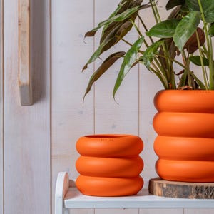 Pode incluir: Dois vasos de cerâmica em laranja vibrante com um design distinto de anéis empilhados e arredondados. O vaso menor está em primeiro plano, enquanto o maior contém uma planta verde e repousa sobre uma base de madeira com acabamento liso.